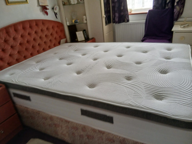 A white tencel cool tufted mattress on a bed frame in a bedroom setting, with a reddish-orange tufted headboard, shelving, and a window in the background
